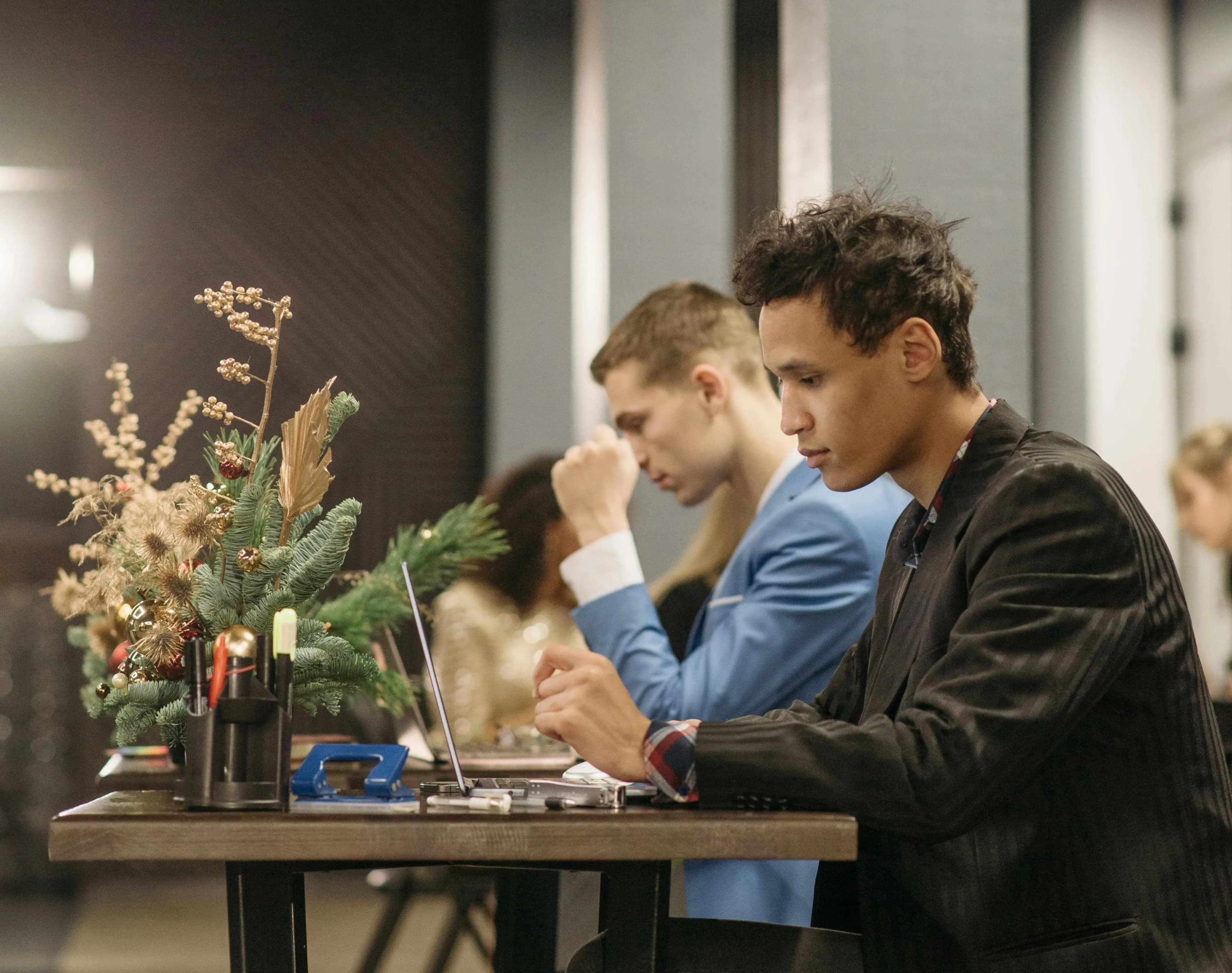 Two men sitting at a table alongside each other, both are working on their laptops