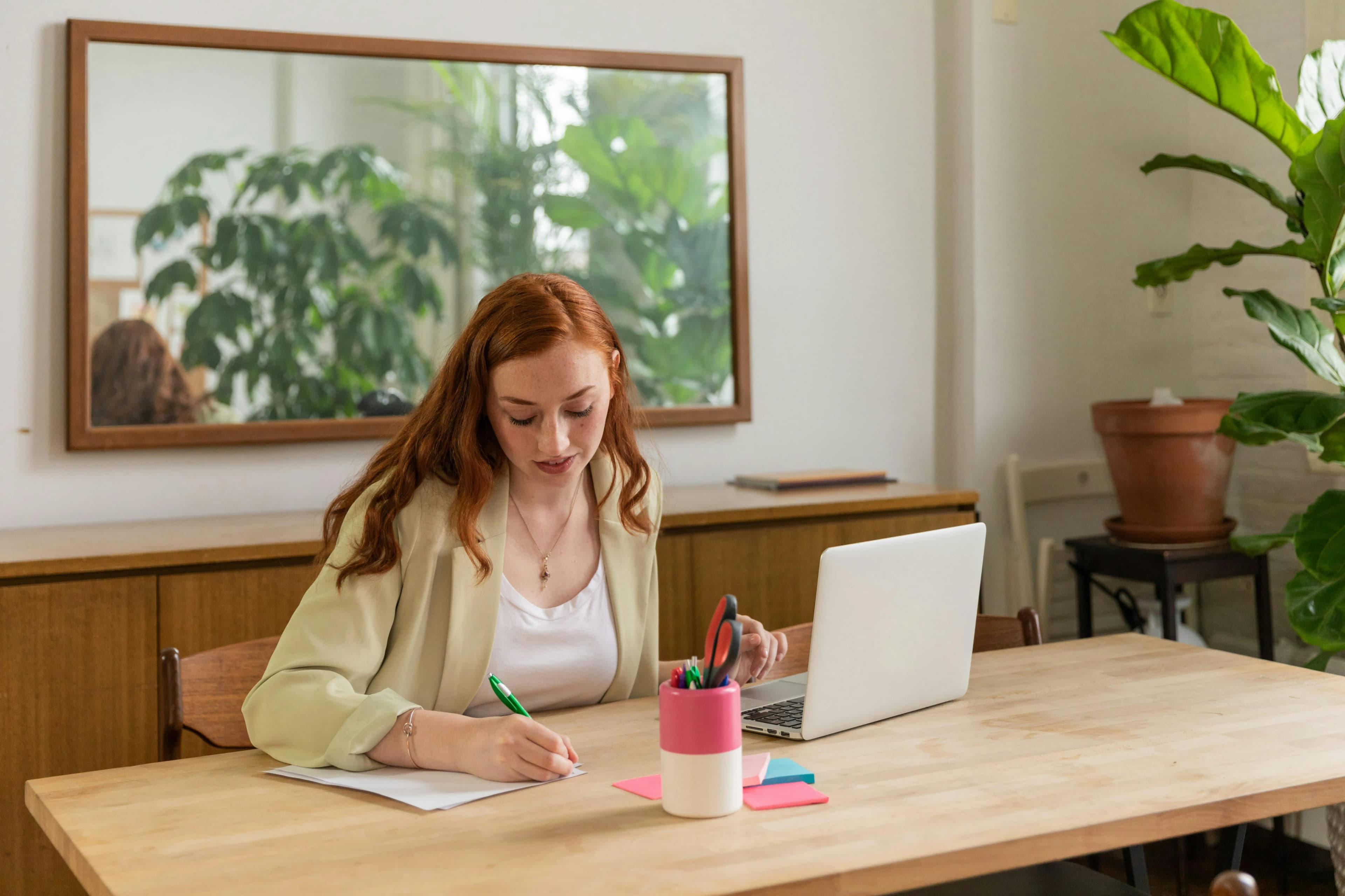 Woman sitting at a table writing on a piece of paper, she also has her laptop open.